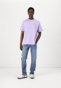 Camiseta de cuello redondo de color lavanda, de corte relajado. Combinada con jeans rectos de tiro medio en azul claro y zapatillas blancas con detalles en negro. Fondo liso.
