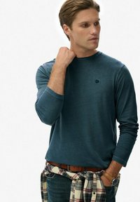 Donker teal lange mouwen shirt met ronde hals, voorzien van een klein logo op de borst. Draagt een geruit shirt knoopt om de taille en denim jeans.