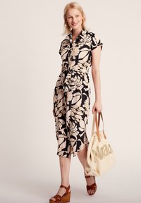 Robe à motifs floraux en navy et crème, manches courtes, taille ceinturée, longueur genou, associée à un sac fourre-tout beige et des sandales compensées marron.
