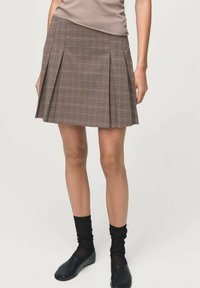 Jupe plissée à carreaux bruns avec une taille ajustée. Texture de tissu douce, longueur mi-cuisse, associée à des chaussettes noires et des chaussures simples.