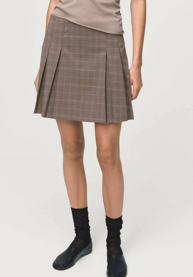 Jupe plissée à carreaux bruns avec une taille ajustée. Texture de tissu douce, longueur mi-cuisse, associée à des chaussettes noires et des chaussures simples.