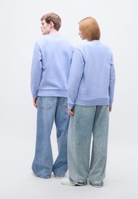 Ανοιχτό μπλε φούτερ με ριμπ τελειώματα φορεμένα πάνω από άνετα, σκισμένα ανοιχτόχρωμα τζιν. Το φόντο είναι μονόχρωμο λευκό.