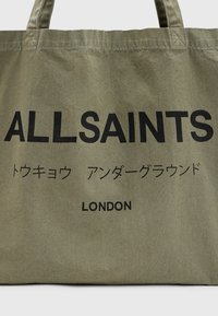 Borsa tote in tela verde oliva con testo nero che legge "ALLSAINTS" e caratteri giapponesi, dotata di manici robusti e finitura opaca.