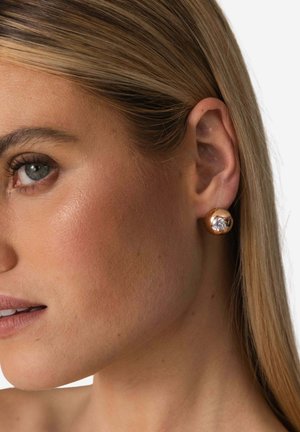 Ohrstecker in Goldoptik mit einem runden, polierten Design, das in der Mitte einen klaren Edelstein zeigt, getragen am Ohr eines Models.