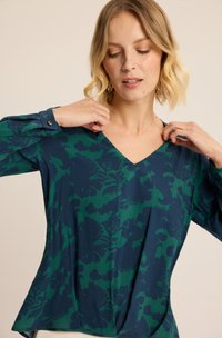 Bluzka z dekoltem w serek w ciemnoniebieskim kolorze z zielonym kwiatowym wzorem, z długimi rękawami i mankietami zapinanymi na guziki; miękki materiał w luźnym kroju.