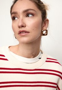 Cremefarbener Pullover mit roten Streifen und geripptem Ausschnitt, getragen über einem weiß-schwarz gestreiften Hemd. Goldener quadratischer Ohrring mit hohlem Zentrum.