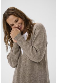 Gaiši brūns adīts kardigans, kas izgatavots no mīksta materiāla ar ribotām tekstūrām, atvērta priekšpuses un garām piedurknēm, valkāts virs baltas blūzes.