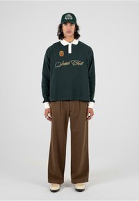 Camisa de manga larga verde oscuro con cuello blanco y texto bordado en dorado que dice "Social Club". Pantalones marrones con pliegues y zapatos beige.