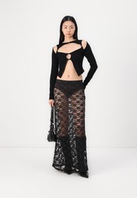Zwart langsleef top met uitsnijdingen en een sleutelgat, gecombineerd met een transparante zwarte kanten rok. Model houdt een kleine zwarte handtas vast.