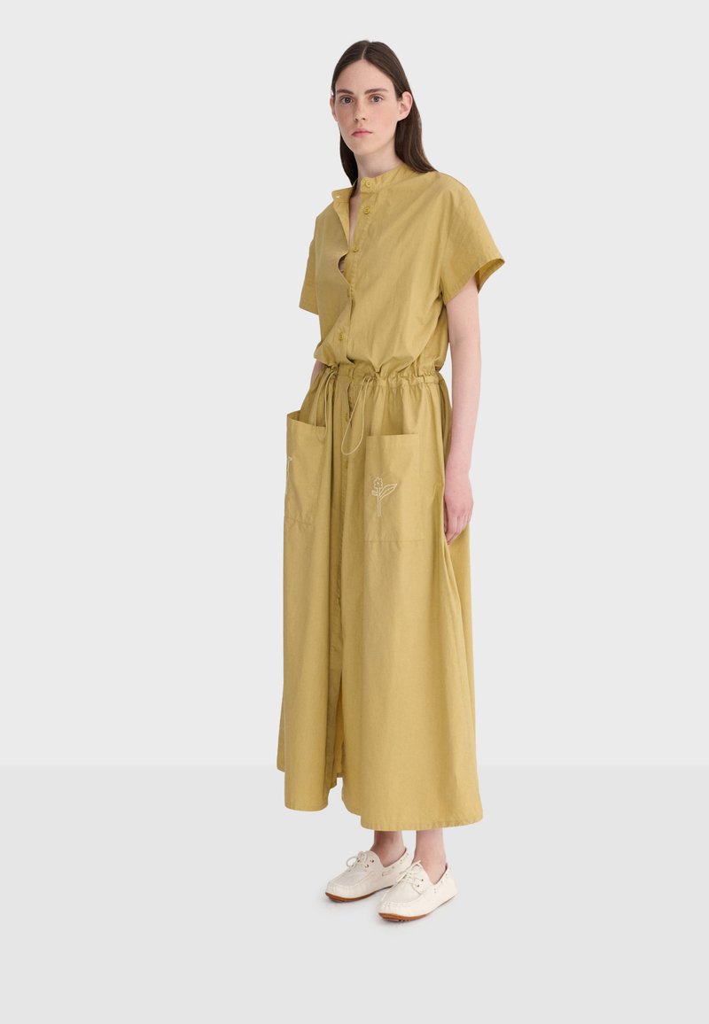 Femme vêtue d'une robe chemise jaune moutarde à manches courtes, taille froncée avec cordon, grandes poches avant, et chaussures blanches décontractées, debout immobile.