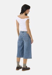 Culotte in denim blu con una vestibilità comoda, dotate di vita alta, gambe larghe e due tasche posteriori. Abbinati a un top bianco off-the-shoulder e scarpe scure.