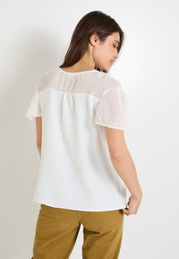 Top blanco con mangas cortas texturizadas y un patrón sutil. El cuerpo principal es liso, con un dobladillo ligeramente curvado.