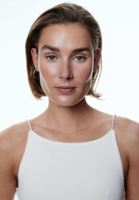 Weißes geripptes Tanktop mit dekorativen transparenten Trägerakzenten. Das Modell hat kurze braune Haare und neutrale Schminke, vor einem schlichten Hintergrund.