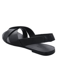 Mustaan nahkakengän sandaali, jossa on ristikkäiset hihnat ja takahihna, litte ä pohja, kuvattuna takakautta valkoista taustaa vasten.