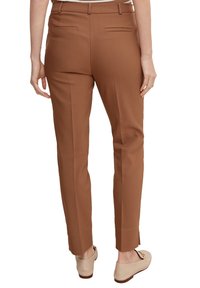 Pantalon marron sur mesure à coupe slim, avec poches arrière et texture lisse, associé à des chaussures de couleur claire sur un fond neutre.