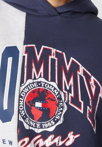 Felpa con cappuccio caratterizzata da un pannello sinistro grigio e un pannello destro blu navy, con la scritta audace "TOMMY" e una grafica di un globo in rosso e bianco.