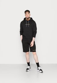 Completo con felpa nera e pantaloni corti di Calvin Klein, dotato di una tasca frontale e logo ricamato. Il modello indossa sneakers bianche e nere.