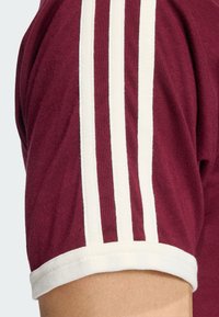 Μπορντό κοντομάνικο πουκάμισο με τρεις λευκές ρίγες στο μανίκι και ριμπ λευκό γιακά. Το υλικό φαίνεται μαλακό και με υφή.