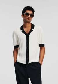 Witte blouse met korte mouwen, zwarte kraag en manchetten, met kabelgebreide structuur en zwarte knopen, gedragen met donkere broek en zonnebril.