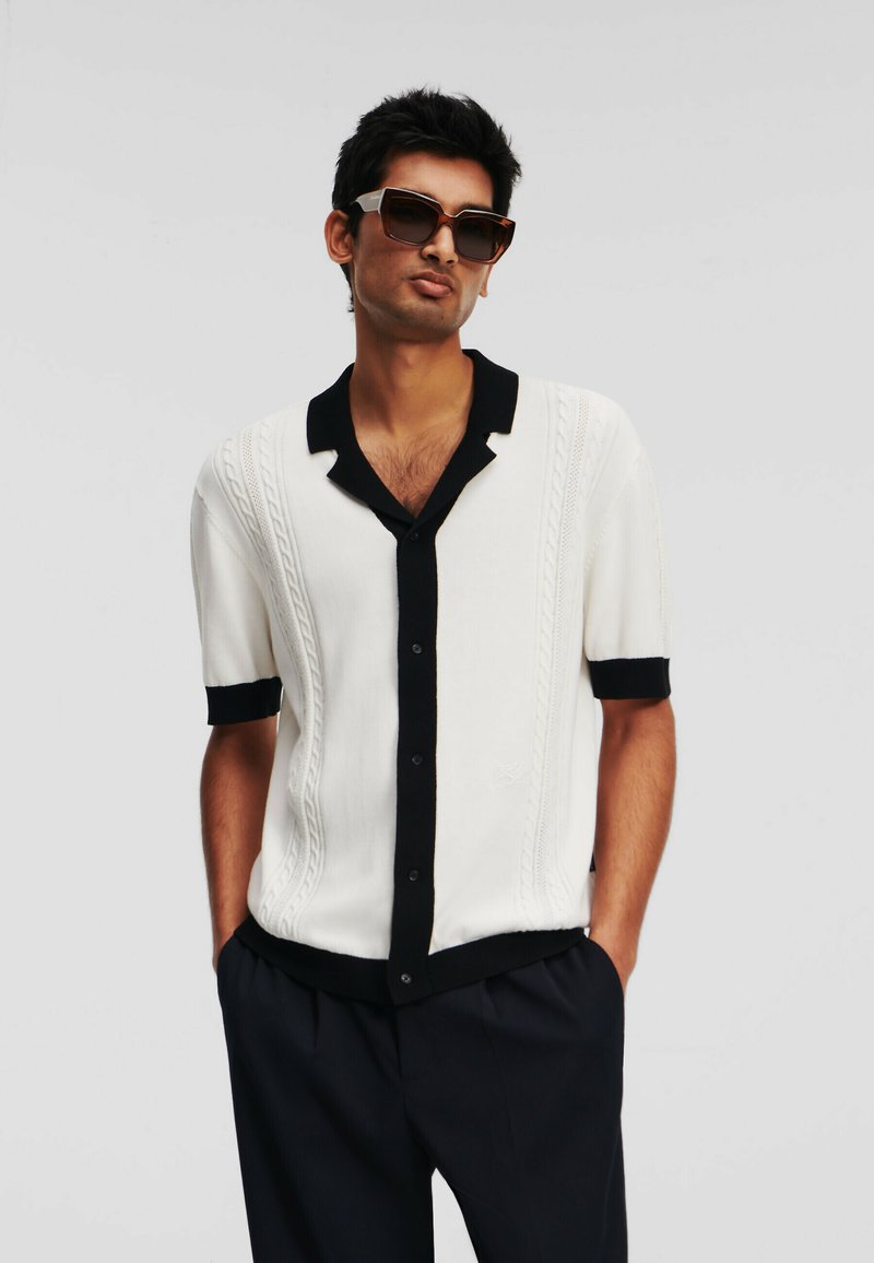 Witte blouse met korte mouwen, zwarte kraag en manchetten, met kabelgebreide structuur en zwarte knopen, gedragen met donkere broek en zonnebril.