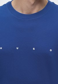 Gros plan d’une personne portant une chemise bleue avec des lettres blanches espacées formant le mot « HUGO » sur la poitrine.