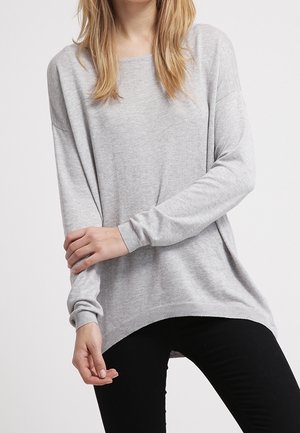 Kobieta w jasnoszarym długim swetrze z dzianiny i czarnych spodniach, stojąca z założonymi rękami na tle jednolitego tła.