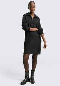 Czarna dzianinowa sukienka z zamkiem przy kołnierzu, długimi rękawami i prążkowaną fakturą. Noszona z czarnymi botkami sięgającymi kostek i przezroczystymi rajstopami.