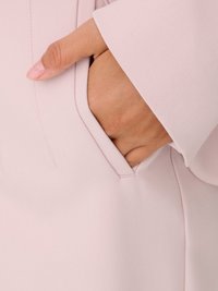 Roka daļēji ielikta gaiši rozā apģērba kabatā ar redzamām šuvēm un gludu auduma tekstūru.