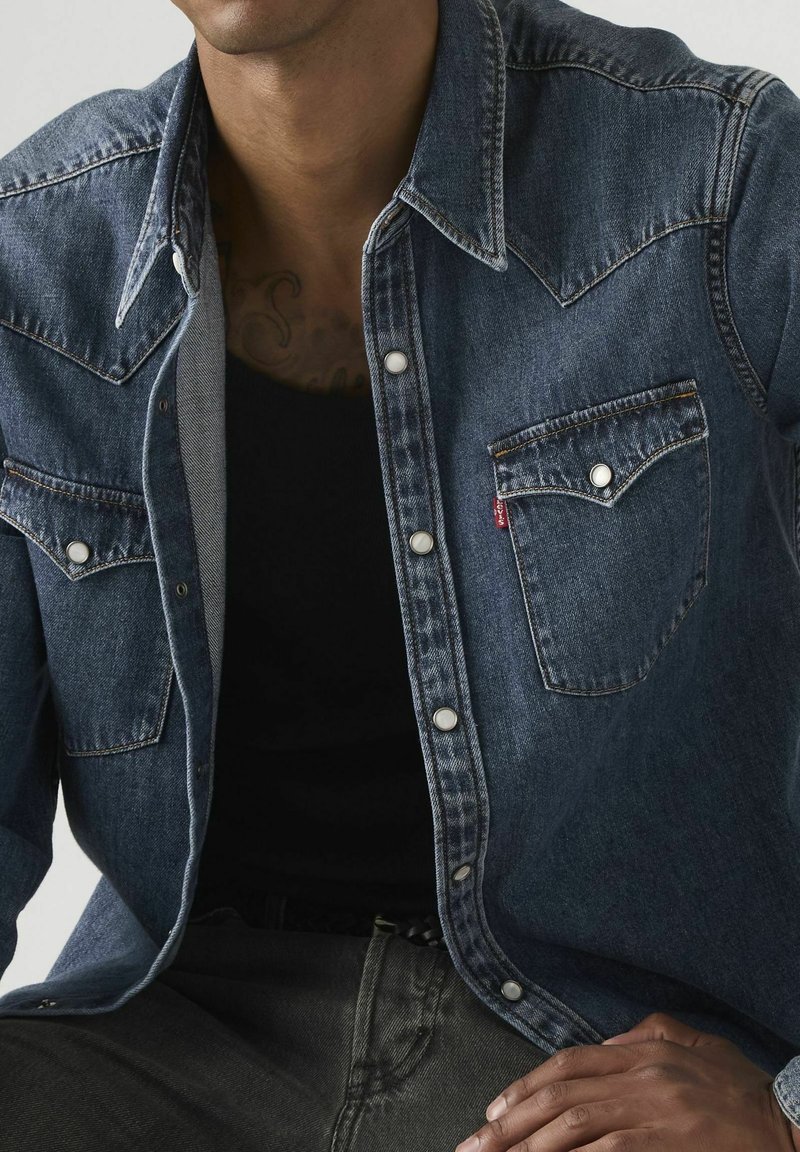 Mann trägt eine dunkelblaue Jeansjacke mit Druckknöpfen über einem schwarzen Oberteil, sitzt und legt eine Hand auf seinen Oberschenkel.