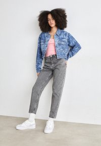 Jeansjacke mit Palmbaum-Muster in Hellblau, pinkes Tanktop, graue hochgeschnittene Jeans und weiße Sneakers mit Plateau-Sohle.