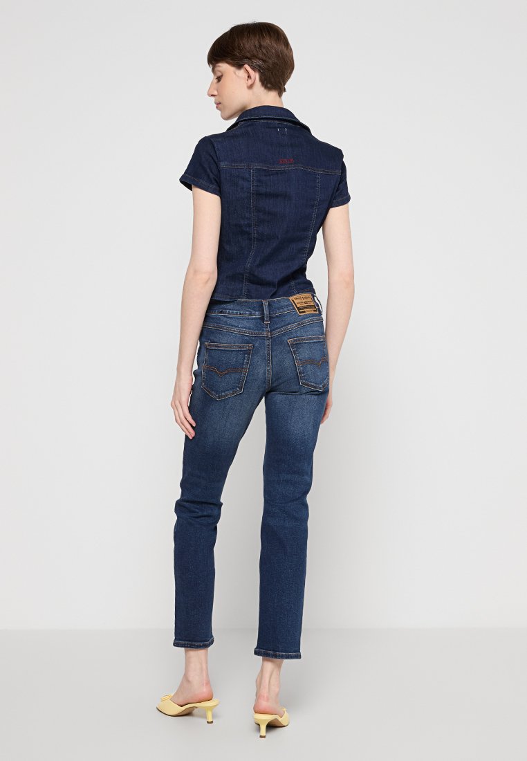 Donkerdenim jumpsuit met korte mouwen, aansluitend ontwerp en een logo-opdruk aan de achterkant. Beschikt over twee achterzakken en een lichte vervaging, gecombineerd met gele hakken.