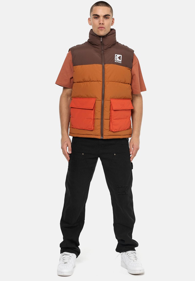 Gevoerde vest in bruin, oranje en roest met hoge kraag, ritssluiting aan de voorkant en twee grote oranje zakken. Draagt over een shirt met korte mouwen.
