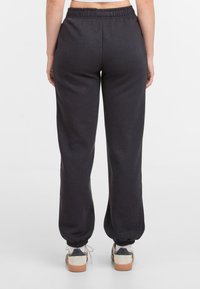 Vrouw in zwarte joggings met elastische taille en witte sneakers, gezien vanaf de achterkant op een plain witte achtergrond.