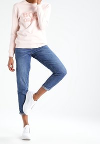 Sudadera rosa claro con el texto "BOSS ORANGE", combinada con jeans de mezclilla azul remangados y zapatillas blancas. Un atuendo simple y casual.