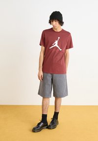 Mørkerød t-shirt med et hvidt grafisk logo, parret med grå denim shorts. Sorte sko med kraftige såler og sorte sokker fuldender looket.