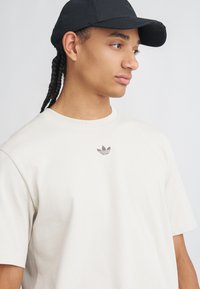 Mladý človek s vrkôčkami, ktorý má na sebe čiernu čiapku a krémové tričko Adidas, pozerá doprava na jednolatom pozadí.