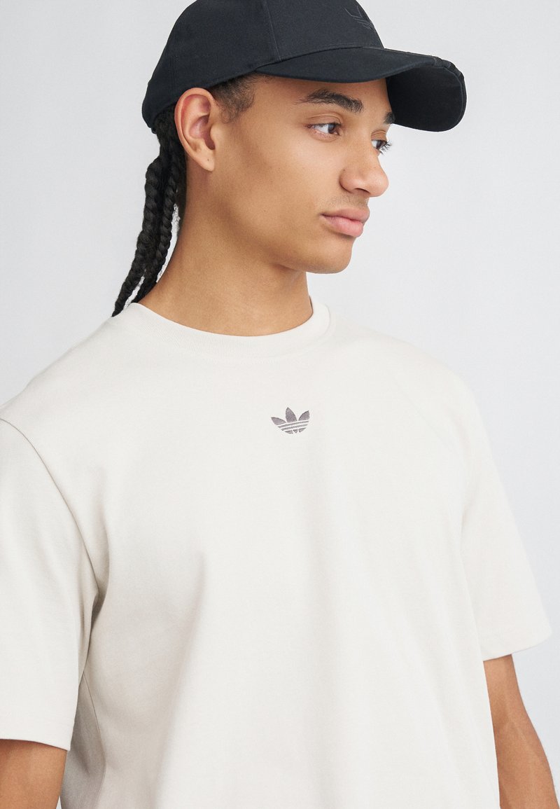 Mladý človek s vrkôčkami, ktorý má na sebe čiernu čiapku a krémové tričko Adidas, pozerá doprava na jednolatom pozadí.