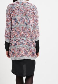 Strickjacke mit einem mehrfarbigen, strukturierten Muster, mit lockeren Ärmeln und einem grauen Saum, die über einem schwarzen Kleidungsstück getragen wird.