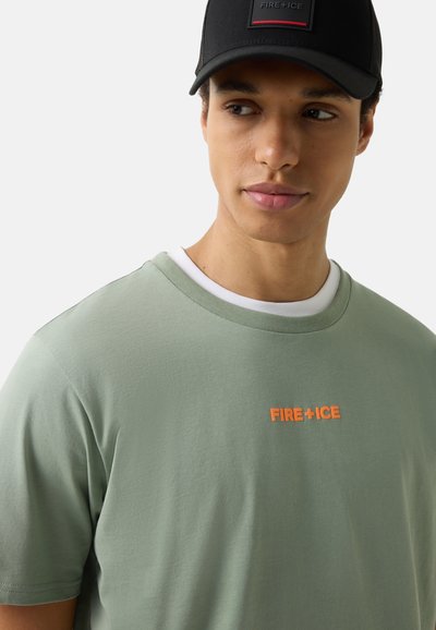 Jeune homme portant un T-shirt vert clair avec le texte orange "FIRE+ICE" et une casquette noire avec le même logo, regardant sur le côté.