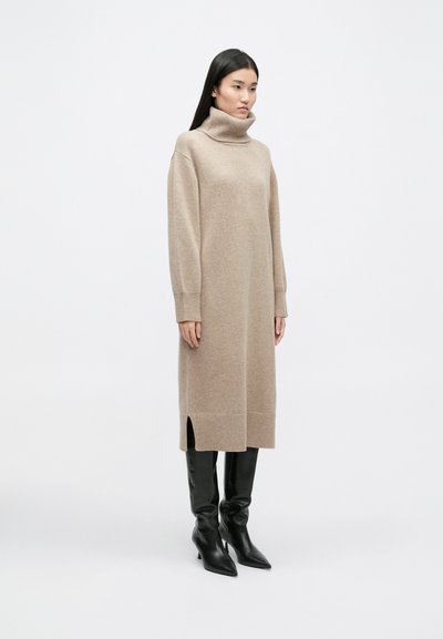 Bež haljina s visokim ovratnikom i dugim rukavima te prorezima sa strane, izrađena od mekanog pletiva, uparena s crnim crnim čizmama do koljena sa špicom.