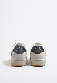 Zapatillas con un cuerpo de ante gris, tapones de talón negros brillantes y acentos blancos. Presentan una marca impresa en el talón y suelas texturizadas.