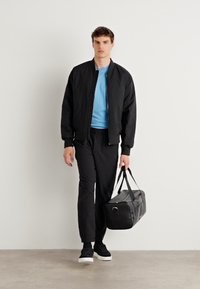 Zwarte bomberjack over een lichtblauw shirt, gecombineerd met zwarte broek en zwarte sneakers, met een zwarte sporttas in de hand. Minimalistisch ontwerp.
