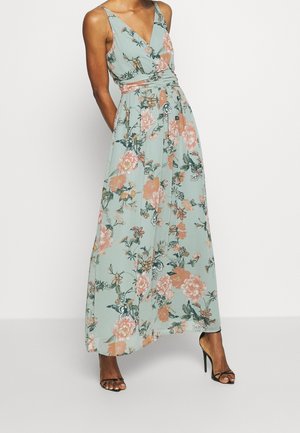 Mujer que lleva un vestido maxi sin mangas, con escote en V, en color verde menta con estampado floral rosa y zapatos de tacón alto negros con tiras, de pie contra un fondo blanco.