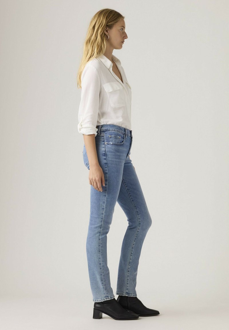 Kvinde stående sidelæns iført en hvid langærmet skjorte, lyseblå jeans og sorte ankelstøvler mod en ensfarvet baggrund.