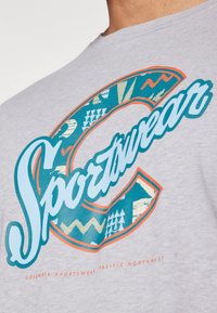 Pilkas marškinėliai su dideliu žalio ir oranžinio atspalvio grafiniu logotipu, kuriame yra įvairių geometrinių raštų ir „Sportswear“ tekstu drąsiai stilizuotu šriftu.