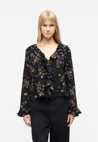 Γυναίκα που φοράει μαύρη μπλούζα με λουλουδάτο σχέδιο, βολάν στο μπροστινό μέρος και στα μανίκια, συνδυασμένη με μαύρο παντελόνι, στέκεται μπροστά σε ένα απλό λευκό φόντο.