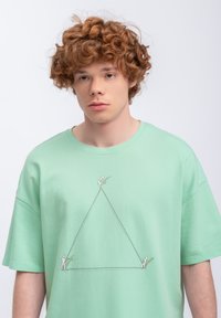 T-shirt verde menta con una stampa grafica di due figure alla base e una alla sommità di un triangolo, contornato da un design semplice.
