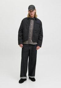 Jeune personne portant une casquette noire, une veste noire, une chemise à carreaux, un large jean sombre avec des revers, et des chaussures noires, se tenant contre un fond uni clair.