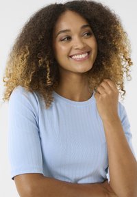 Top de rayas azul claro con mangas cortas, que presenta un escote redondo. La modelo tiene el cabello rizado y sostiene suavemente su mano sobre el hombro.