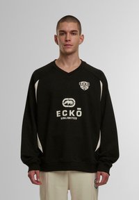 Juodas džemperis ilgomis rankovėmis, su V formos iškirpte ir baltais akcentais. Pasipuošęs logotipais ant krūtinės ir peties. Lygus audinys, kasdieniškas kirpimas.